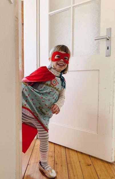 A 4-year-old child dressed up in a cape and mask, standing in a playroom. The child has a determined expression and is holding a toy sword, ready for an imaginary adventure.