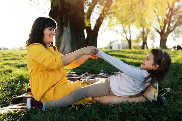 A mother kneeling down to talk eye-to-eye with her son, holding his hands in a gesture of reassurance and support. They are in a park-like setting with trees in the background.