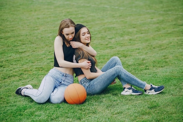A smiling mother kneeling next to her son. They are sitting outside in late afternoon. Both are smiling brightly. He is holding a soccer ball.