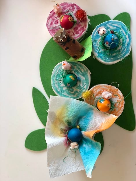 A close-up shot of a colorful nature art collage created by a child. The collage includes pressed leaves, dried flowers, small twigs, and sand, arranged on a piece of sturdy paper.