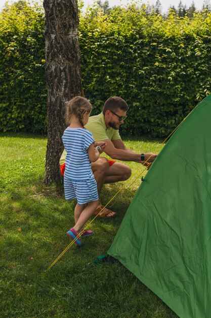 A young boy and girl are enthusiastically setting up a small tent in their backyard, with adult supervision. Camping gear is spread around them, including sleeping bags and backpacks. The scene is set during the daytime.