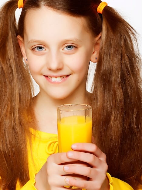 A close-up of a glass filled with a vibrant orange and yellow smoothie, garnished with a slice of orange on the rim. The background shows a child smiling, holding a matching smoothie.