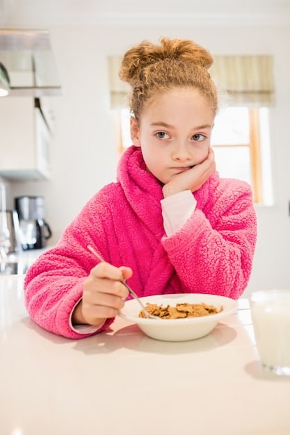 Close-up shot of a child happily eating a bowl of oatmeal topped with fresh berries and a sprinkle of nuts. The background is a blurred kitchen setting.