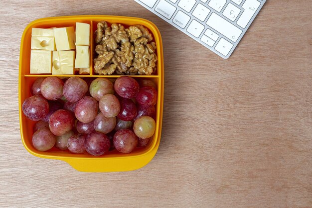 A top-down view of a divided snack container filled with various healthy options, including grapes, cheese cubes, whole-grain crackers, and cherry tomatoes. The container is placed on a colorful, child-friendly placemat.