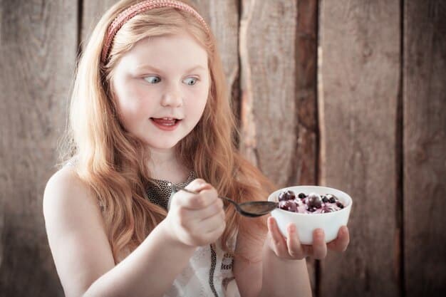 A child sitting at a table, happily eating a bowl of yogurt topped with fresh berries and a sprinkle of granola. The child is smiling and holding a spoon.