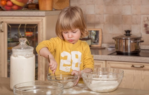 A child, around 6 years old, is carefully pouring pre-measured ingredients into a mixing bowl while an adult guides their hand. The child is wearing a bright apron and has a focused expression, highlighting the learning and collaboration aspect of cooking together.