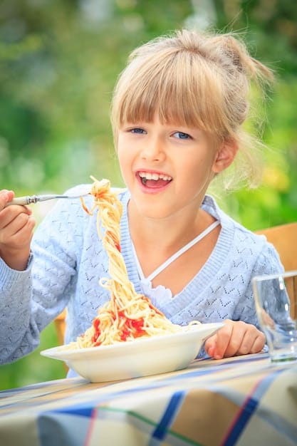 A young girl smiling while eating a colorful plate of plant-based pasta with vegetables. She is sitting at a kitchen table with a bright and cheerful background.