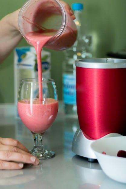 A close-up shot of a colorful smoothie being poured from a blender into a clear sippy cup, garnished with a few fresh berries on the side. The smoothie's vibrant green color indicates the inclusion of vegetables. The background is a modern kitchen counter with a few healthy ingredients like spinach and bananas visible.