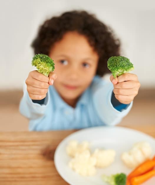 A baby happily reaching for and grabbing a piece of soft-cooked broccoli from a plate in front of them, with a few other finger foods like avocado slices and cooked sweet potato on the plate.