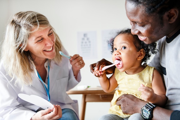 A diverse group of parents smiling and interacting during a pediatrician appointment. The doctor is gently examining a baby, and a chart about Vitamin D is visible in the background.