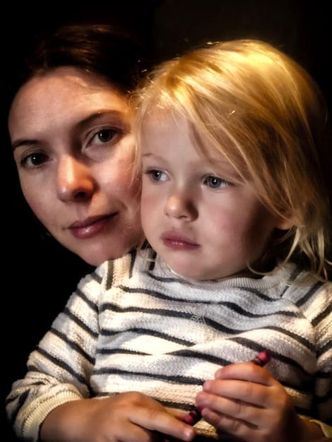 A close-up shot of a baby's face expressing distress as the mother walks away, capturing the raw emotion of separation anxiety. The setting is a brightly colored nursery.